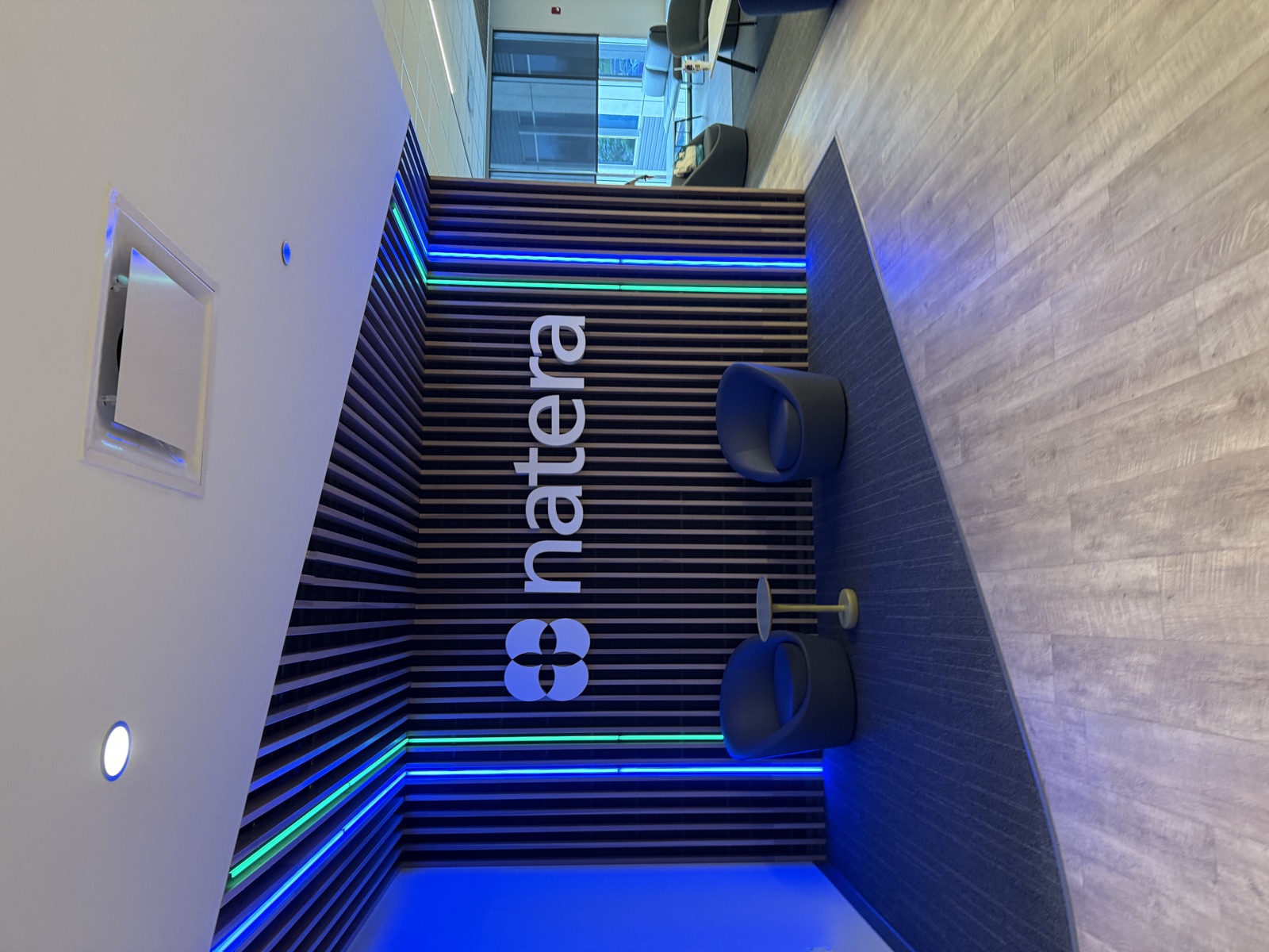 My new home base, when I'm not at home. The Natera office lobby. The company name and logo, a clover-like cluster of four overlapping circles, are mounted in white on a feature wall made of vertical wooden slats. Two vertical strips of blue and green LED lighting flank either side of the wall, with the same colored light continuing across the slatted ceiling overhead. Two round blue swivel chairs sit on a dark gray rug in front of the wall, separated by a small mustard-yellow side table. Beyond the lobby, a glimpse of an adjacent room with more lounge seating and a window onto another office building.