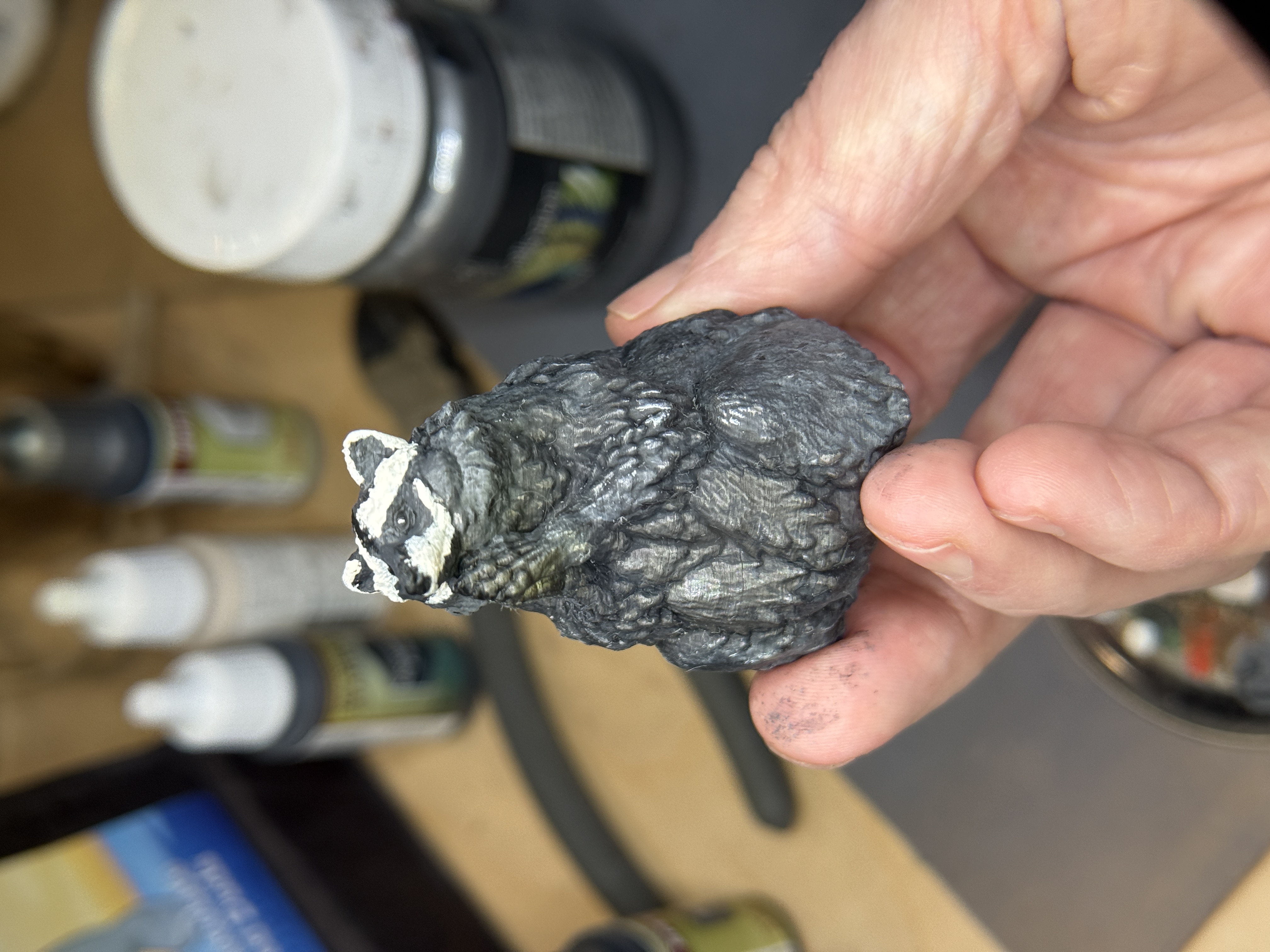 This little guy was printed and painted to be hidden at the con. He didn't last the first evening. A small hand-painted 3D-printed raccoon figurine with detailed gray and white fur, held between two fingers