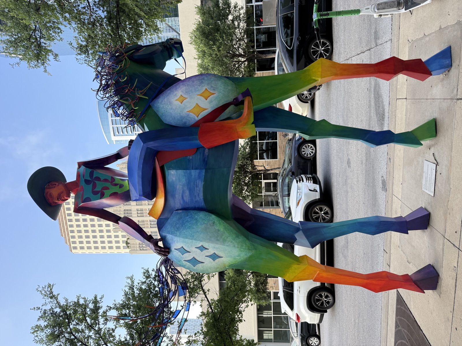A monumental sculpture of a cowboy on horseback, maybe twelve feet tall, standing on a downtown Austin sidewalk. Both horse and rider are rendered in flat geometric panels painted in a rainbow gradient that runs from orange through yellow, green, blue, and purple along their bodies and legs. Pale starbursts decorate the horse's flanks. The horse's mane and tail are made of tangles of brightly colored translucent wire, exploding outward like fireworks. The rider wears a wide black hat and sits stiffly upright with one hand resting behind him on the horse's back. Behind the sculpture, parked cars and trees line the street, and several downtown office buildings rise into a clear blue sky.