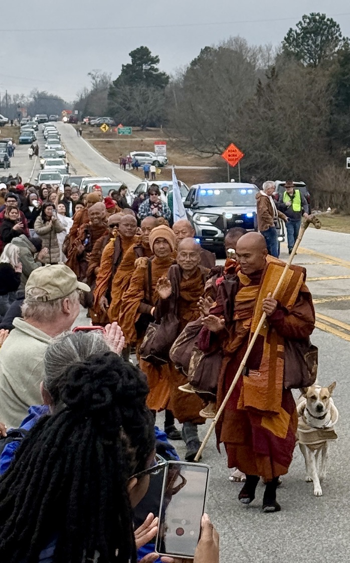Monks in saffron and maroon robes walk down a rural road, led by a monk with a tall walking staff. Aloka the rescue Peace Dog trots alongside. Community members line both sides of the road, many with palms pressed together in greeting, as a police vehicle with blue lights escorts the procession.