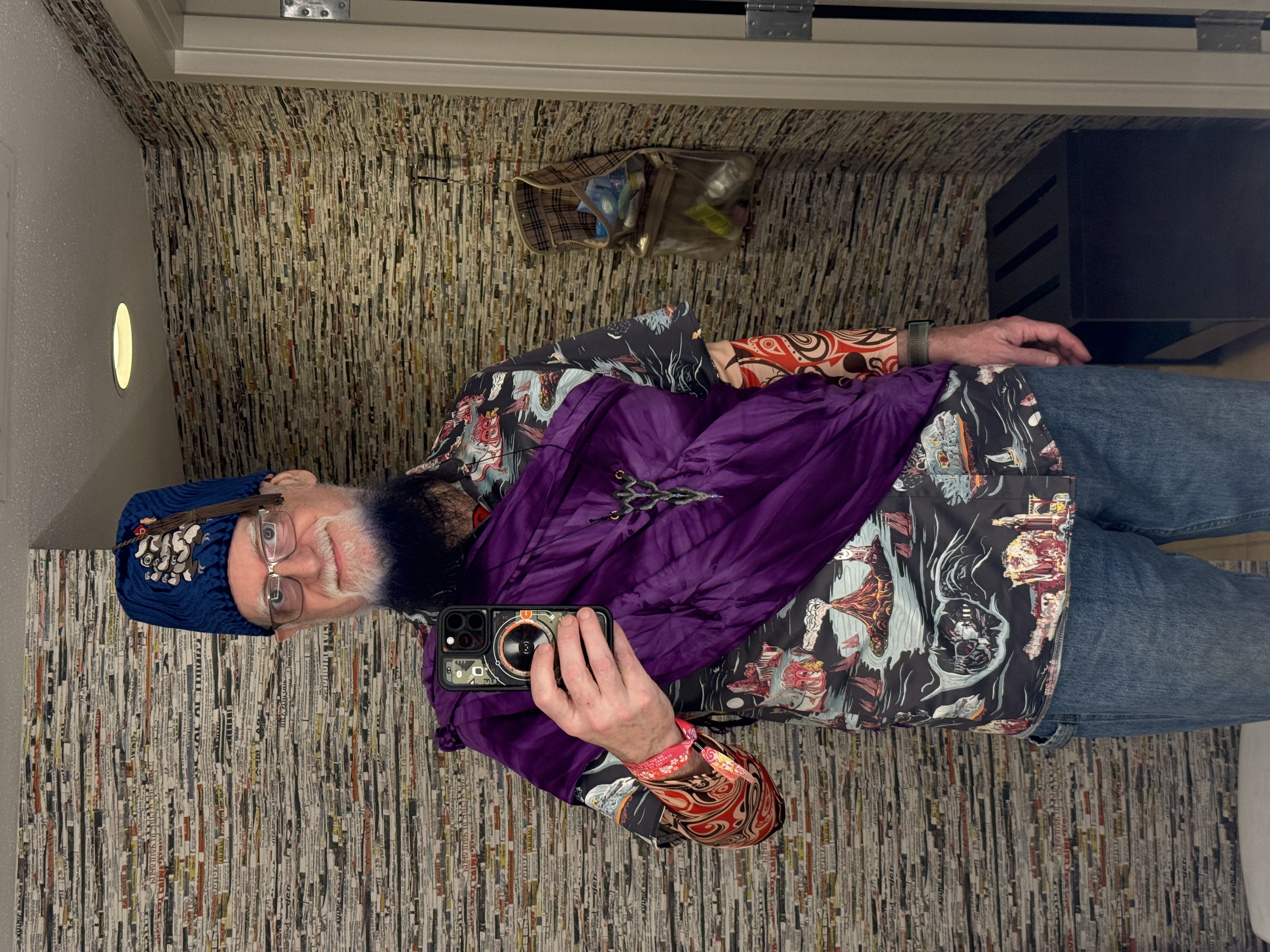Mirror selfie showing Eric in tiki toga party attire: blue embroidered fez, purple sash draped as a toga over a tropical shirt, and the cormorant pendant