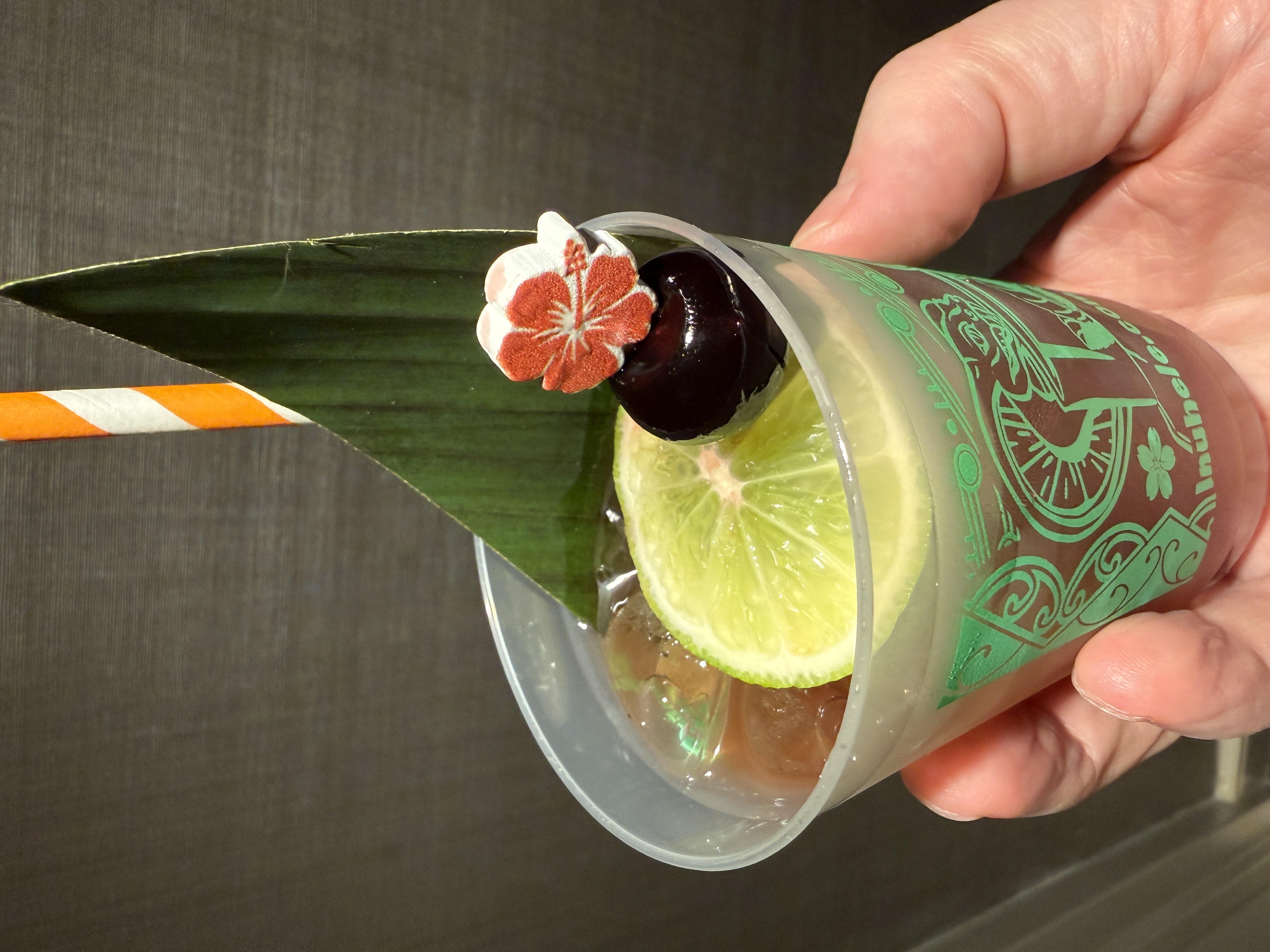 Hand holding a cocktail garnished with a lime wheel, cherry, ti leaf, and small hibiscus pick in an Inuhele-branded cup