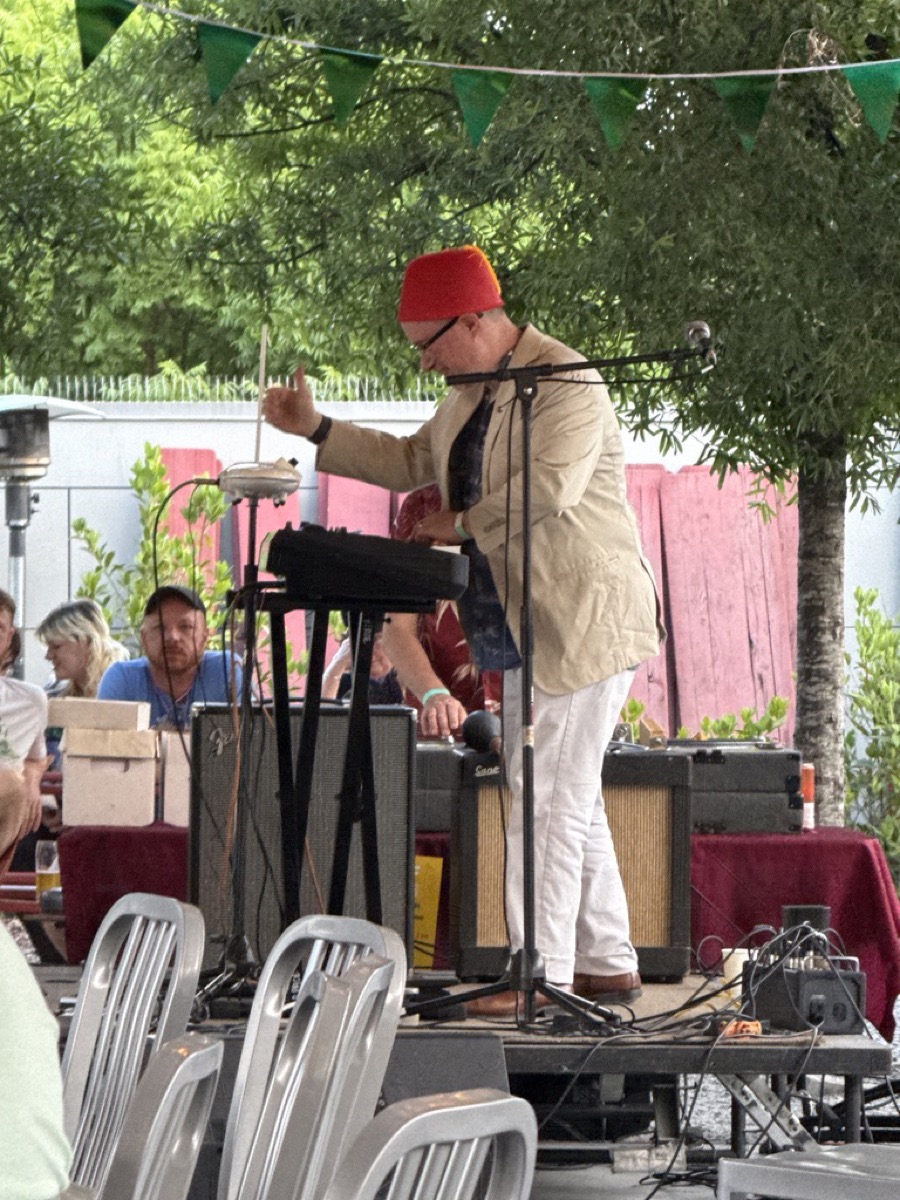 An outdoor stage under green pennant bunting and leafy trees. A performer in a red fez, glasses, a tan jacket, and white pants stands in profile, one hand raised to play a theremin, with vintage Fender amplifiers behind him. Stacked grey folding chairs and a few seated listeners fill the foreground.