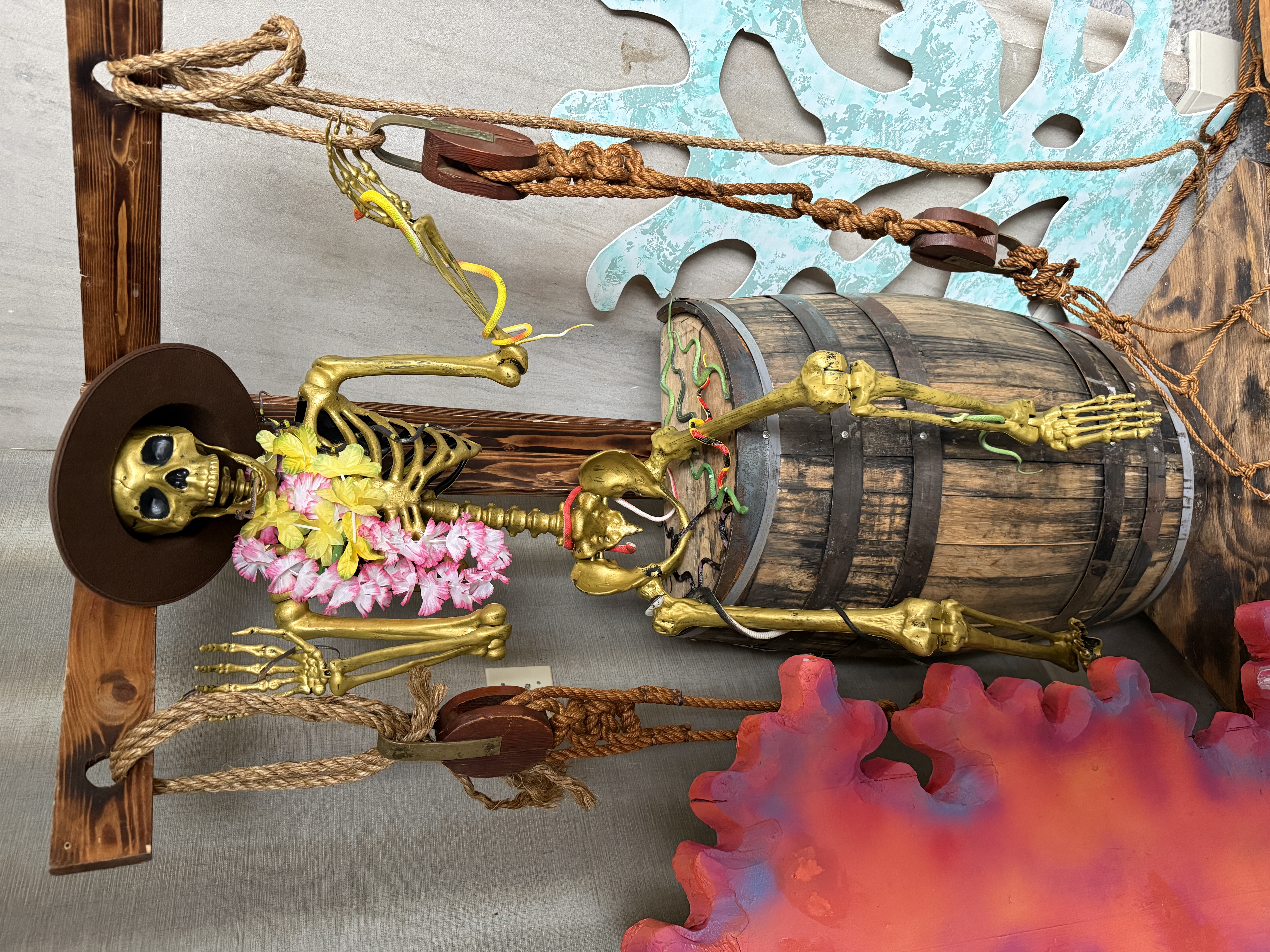 Golden skeleton wearing a hat and lei, seated on a wooden barrel surrounded by ropes and nautical decor