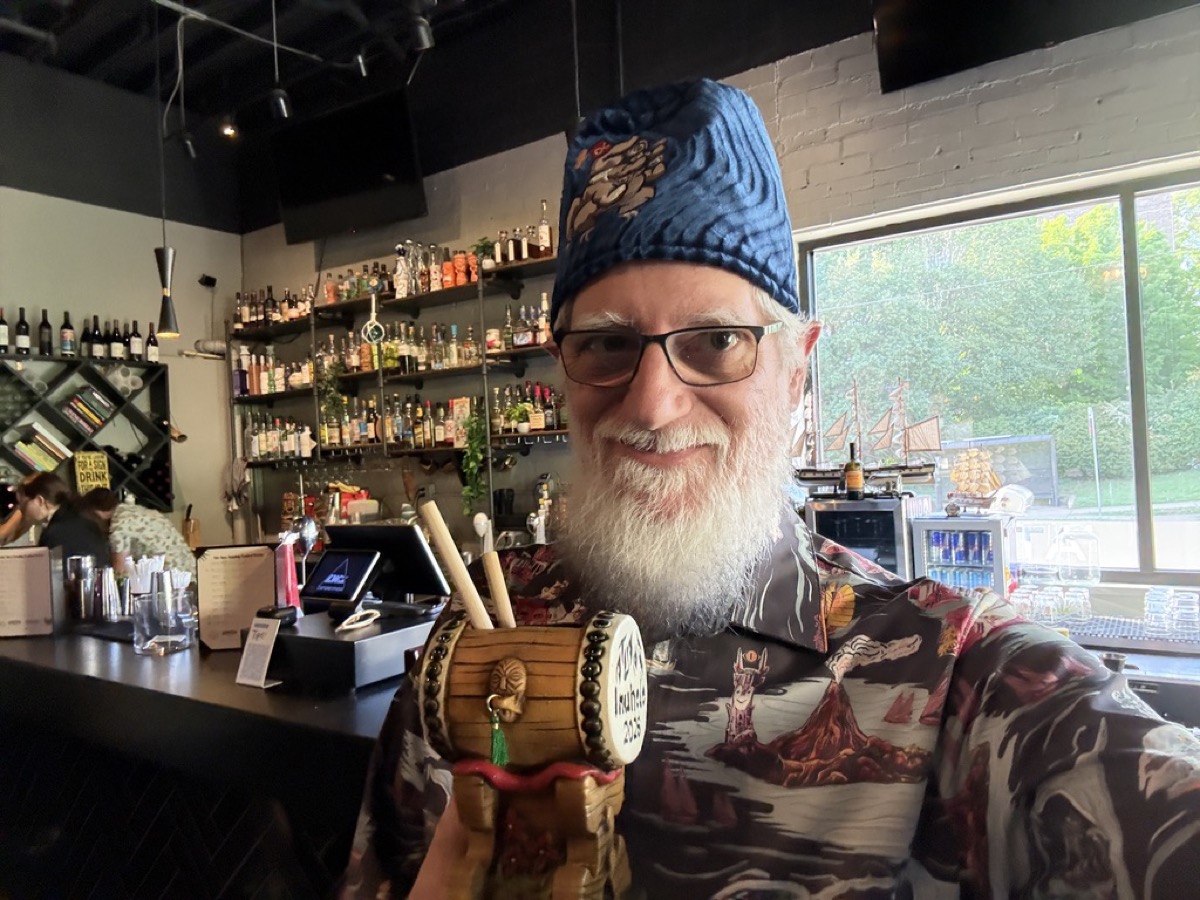 Eric, a bearded man in glasses, smiles in a selfie, wearing a blue patterned fez and a dark Hawaiian shirt printed with volcanoes and sailing ships. He holds up a tiki mug shaped like a small wooden cask with light stir sticks poking out. Behind him is a dimly lit bar lined with shelves of liquor bottles and a model sailing ship on the counter.