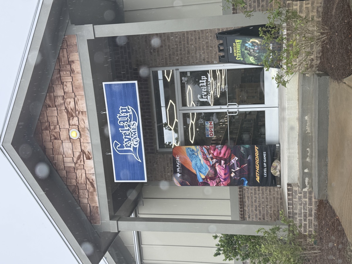 The covered brick entrance of a game store on a rainy day. A blue sign overhead reads 'Level-Up Games' in white lettering, and two glass doors sit under hexagonal ceiling lights, flanked by tall Magic: The Gathering banners on the front steps. Rain spots speckle the lens.