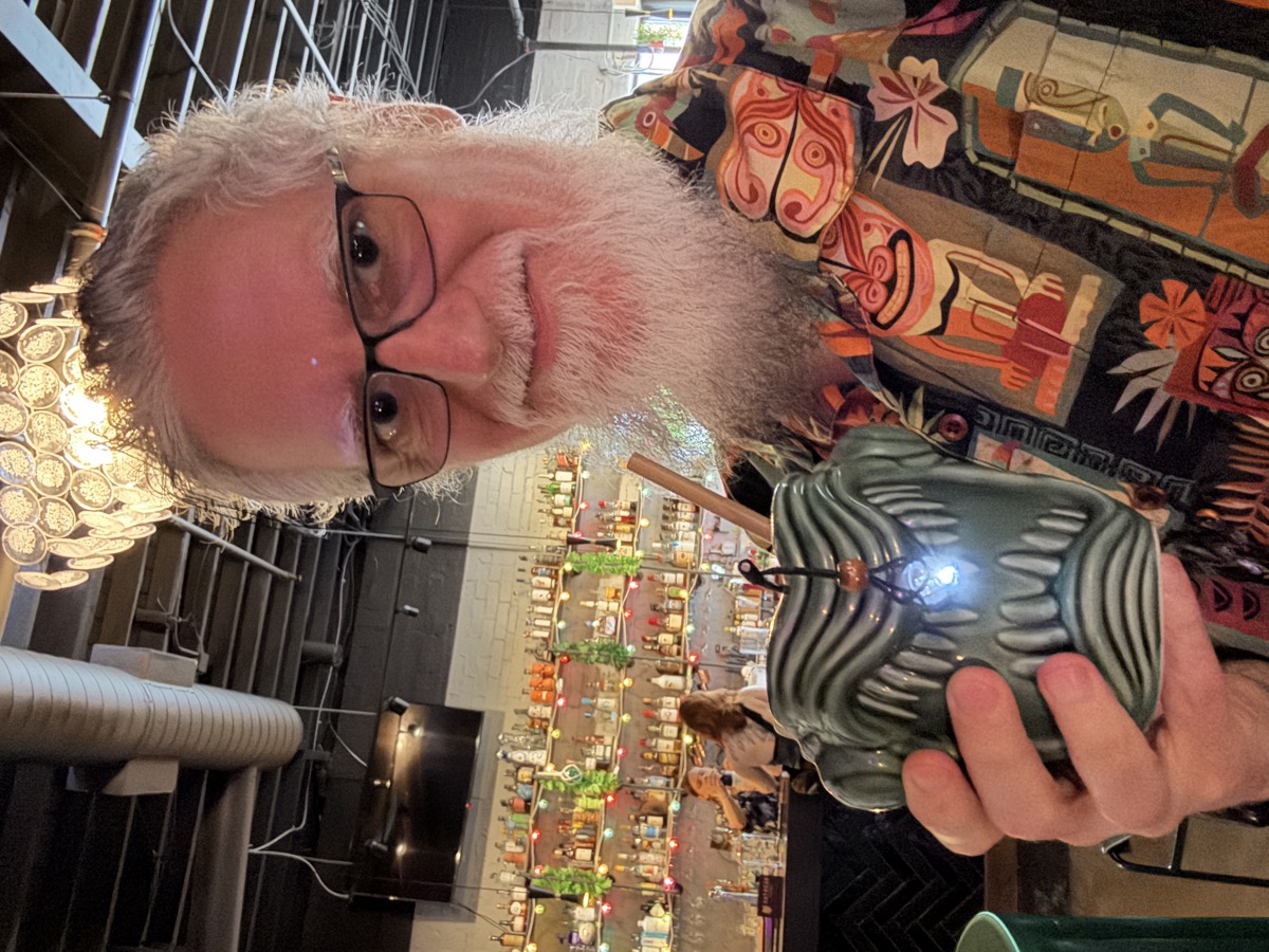 Eric, a bearded man in glasses and a colorful tiki-print shirt, smiles as he holds up a green ceramic mug shaped like a snarling anglerfish, its toothy mouth open and a small lit lure dangling on a stalk out front, a wooden straw poking from the top. Behind him a dim bar is lined with shelves of liquor bottles, strings of colored lights, and a glowing cluster of round lamps overhead.