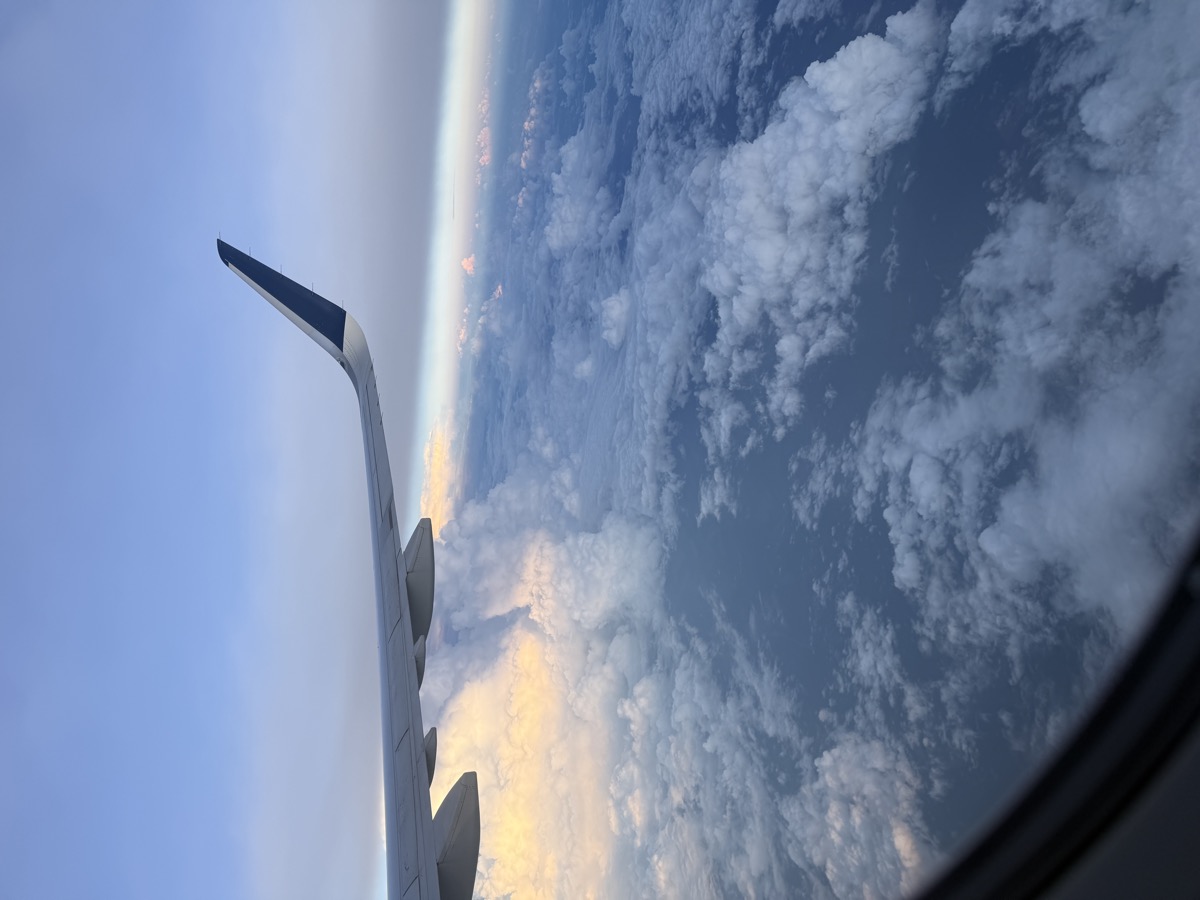 The view from an airplane window seat. A wing with an upturned blue-and-white winglet reaches over a thick floor of clouds, a band of pale gold light along the horizon beneath a clear blue sky. The dark rounded edge of the window frames the lower corners.