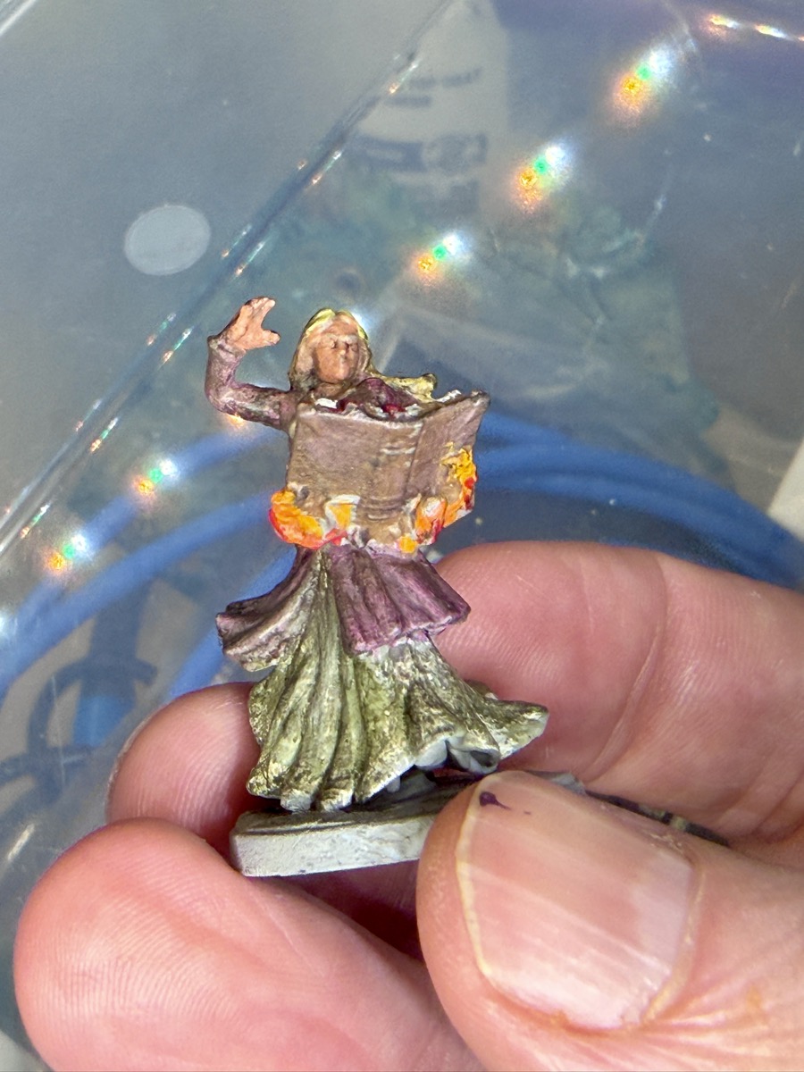 A small plastic tabletop miniature held between finger and thumb. The figure is a robed woman with long blond hair and one hand raised, holding open a large book wreathed in orange and yellow flames. Her robes are painted deep purple over a pale green underskirt. A blue blur fills the out-of-focus background.