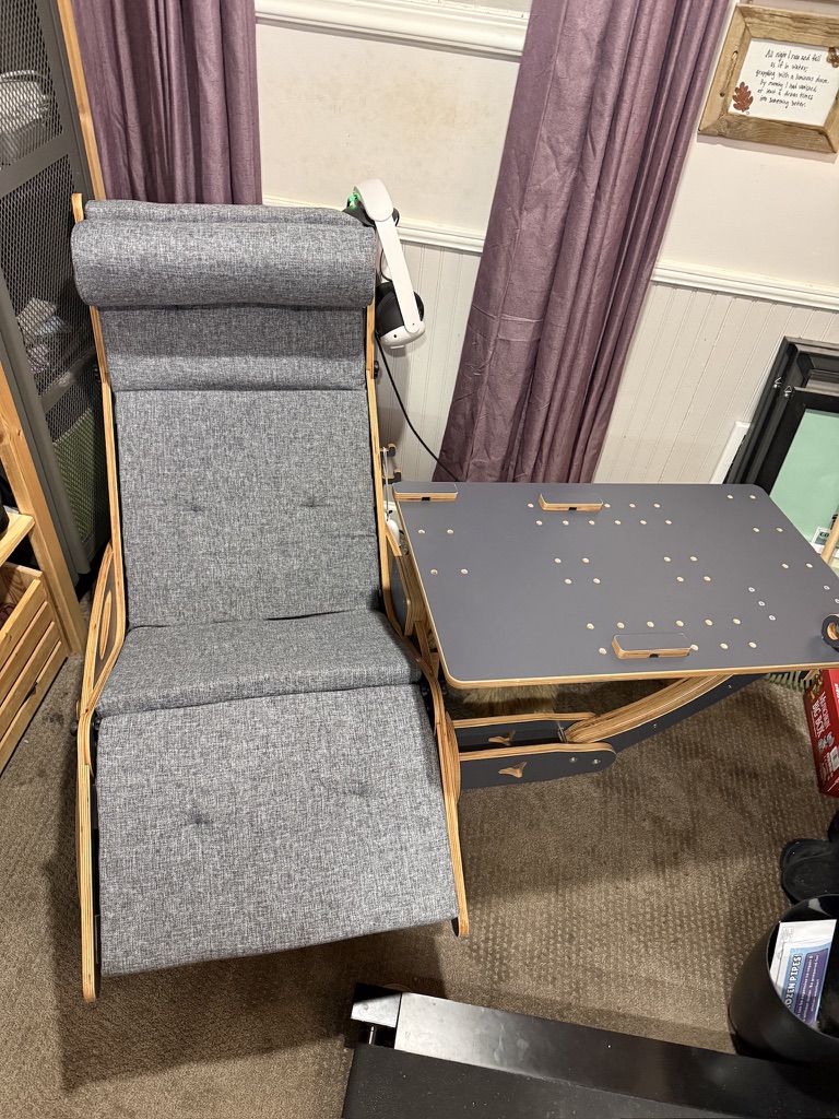 A reclining chair with gray upholstery and a bentwood frame, paired with a dark desk surface with pegboard holes for accessories