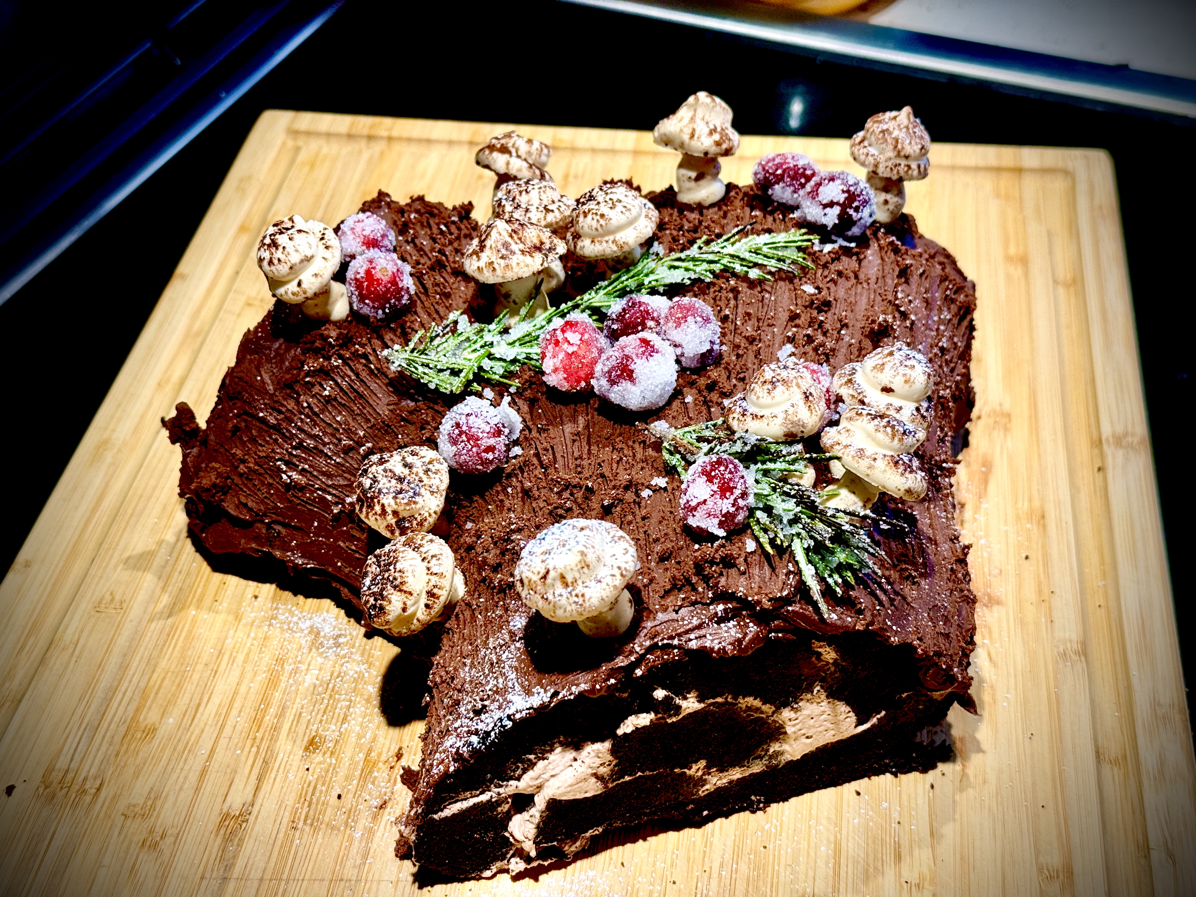 Overhead view of completed yule log cake decorated with meringue mushrooms, sugared cranberries, and sugared rosemary on a wooden cutting board.