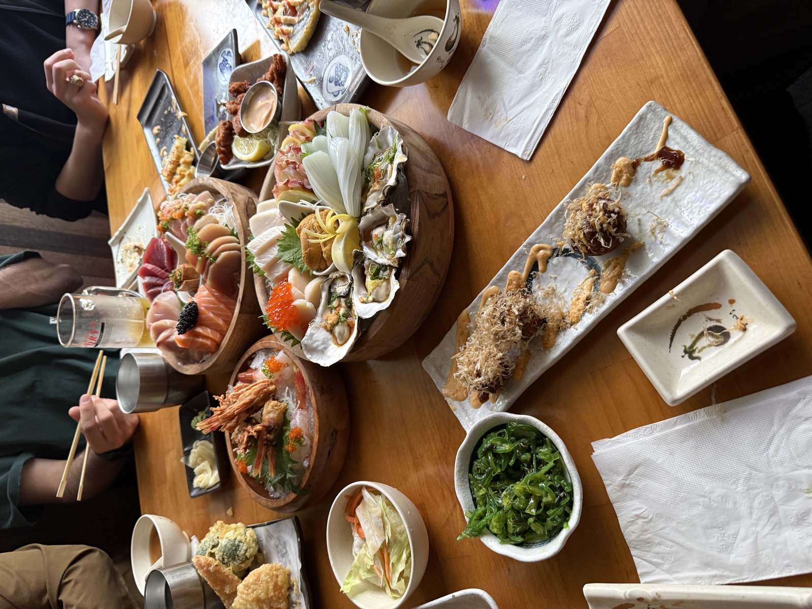A table covered with wooden platters of sashimi, oysters, shrimp, takoyaki, seaweed salad, and tempura at a sushi restaurant in San Mateo