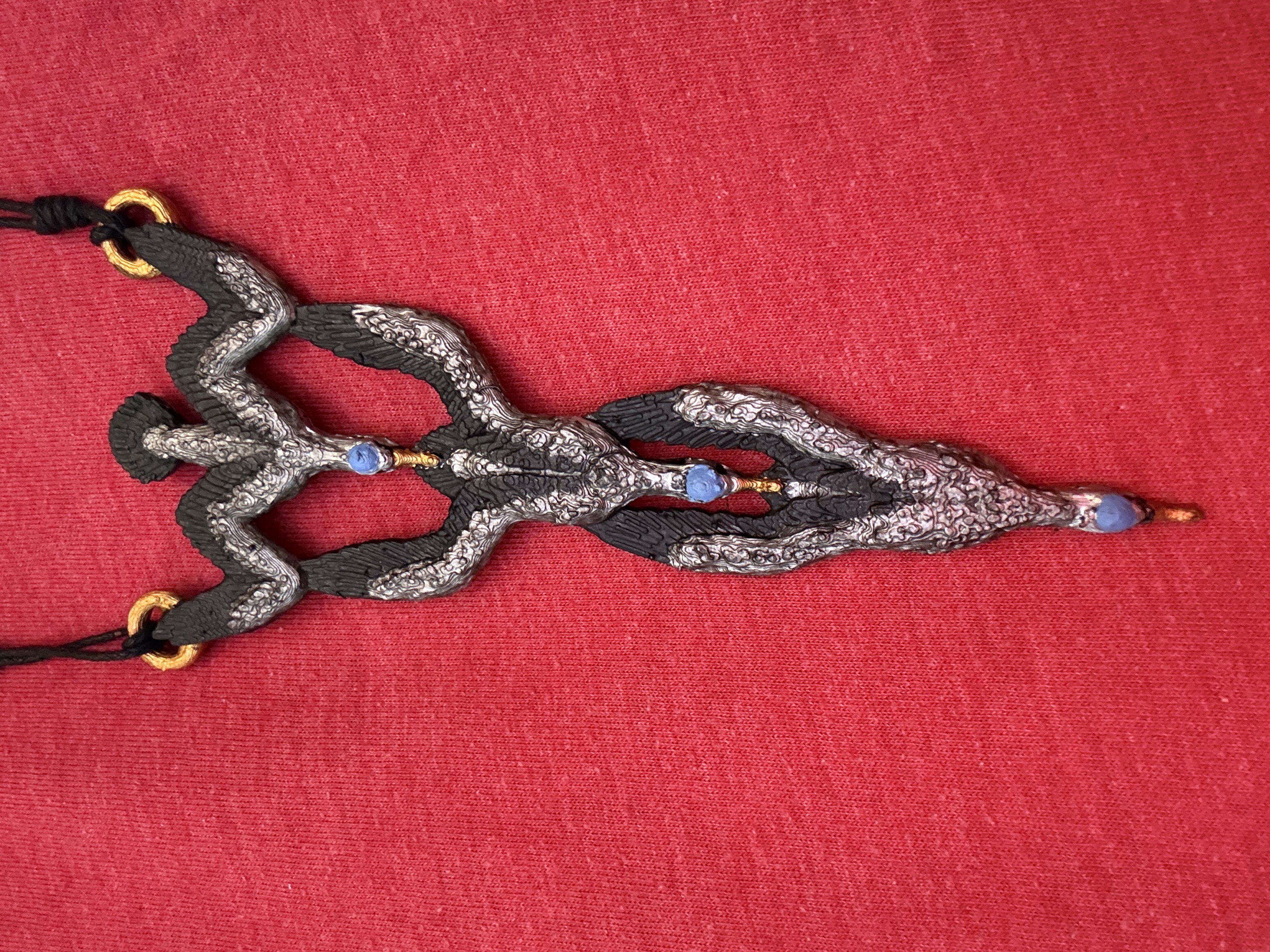 Close-up of a 3D printed cormorant pendant, hand-painted in black and silver with blue and gold accents, hanging on a black cord against a red shirt