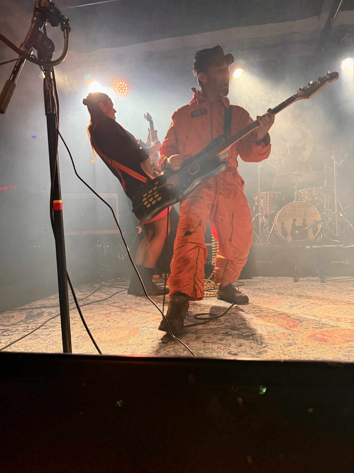 Coco and Avona Nova, somewhere in the haze. Coco in his orange flight suit playing bass in profile while bandmate Avona Nova leans back mid-performance, both lit by stage haze and colored light.