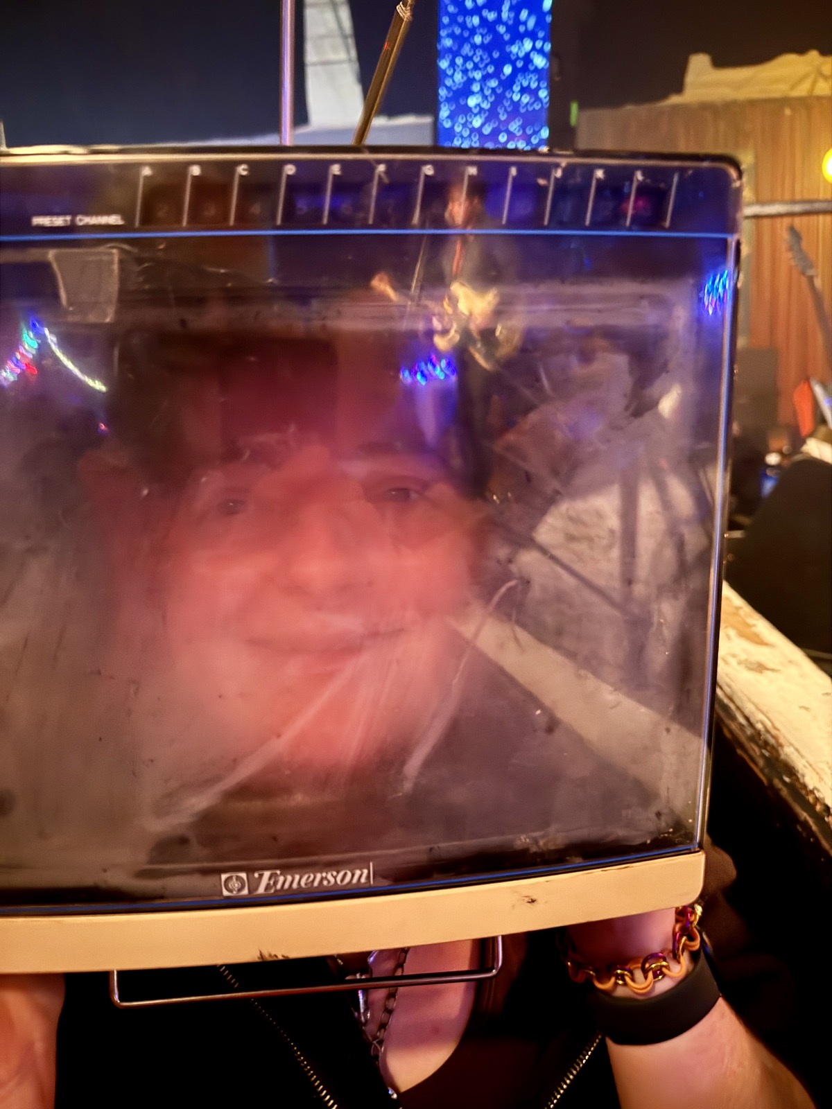 Juniper, broadcasting live from inside an Emerson. A face seen through the screen of a vintage Emerson television set worn as a helmet, lit pink and blue by the stage lights.