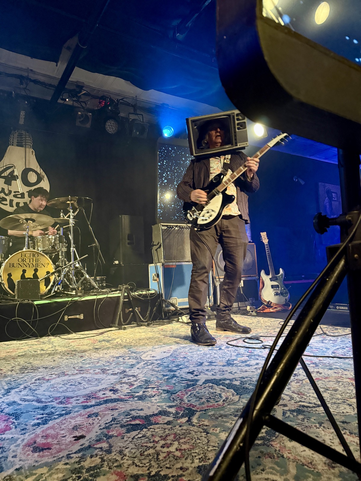 Peter Buck, sitting in. TV helmet and all. A guitarist wearing a vintage television set over his head plays a Rickenbacker on a rug-covered stage, drummer behind him and the 40 Watt light-bulb logo on the wall.