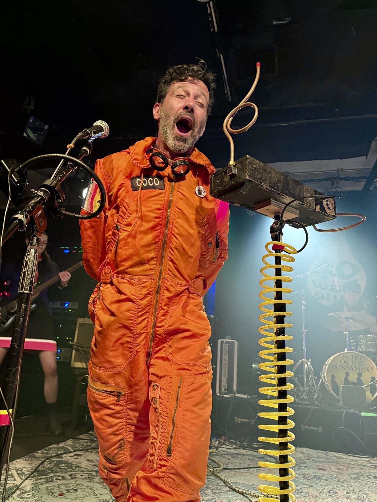 Coco playing the theremin with his face. This is a thing that happened and I have proof. Coco of Man or Astro-man? in an orange flight suit with a COCO name patch and goggles, mouth wide open as he plays a theremin with his face beside a coil of yellow cable on stage at the 40 Watt.