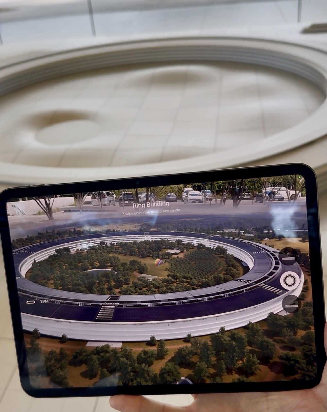 A hand holds a tablet at arm's length. Its screen shows a detailed 3D aerial render of a vast circular glass building ringed by trees, labeled "Ring Building," with onscreen controls and a prompt to swipe up. Behind the tablet, blurred and out of focus, is a large white physical scale model of the same campus.