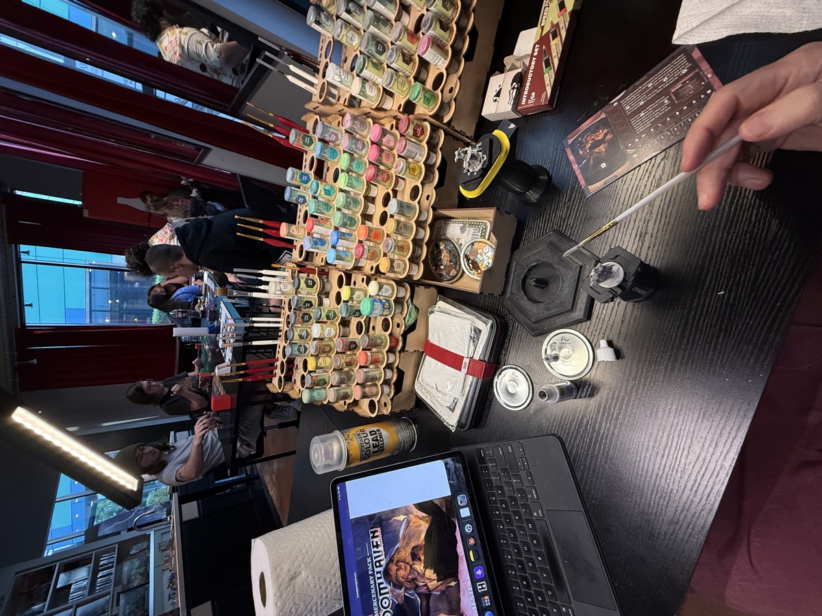 My own paint racks travel well, so I packed them up off my workbench and set up shop at The Painted Heretic for the evening. A painting table at a game store, seen from the painter's seat. An iPad propped on a keyboard case shows reference art for the Gloomhaven mercenary Hail. Tiered wooden racks hold dozens of small paint bottles, a can of Citadel spray primer, and rows of red-handled brushes. A primed miniature on a holder and a character card sit nearby, and a hand holds a fine brush in the foreground. Behind the table, other people paint and play beside tall windows with red curtains, dusk outside.