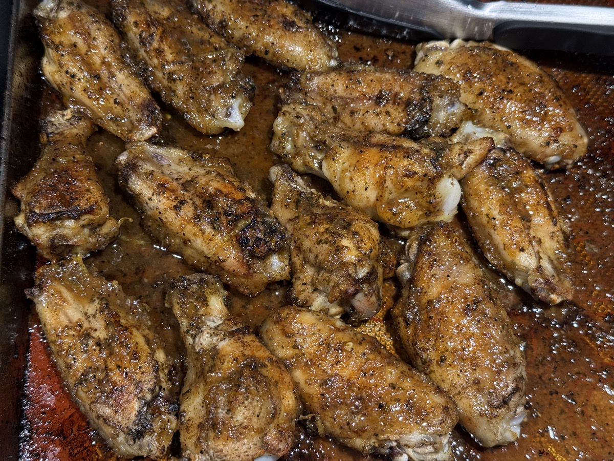 These are my standby whenever wings drop to a good price. This panful was for eating now, with a second batch of raw ones tucked into the freezer for another day. A sheet pan crowded with roasted chicken wings, browned and crisping at the edges, coated in dark herbs and spice and sitting in a shallow pool of their own rendered juices. A metal pan handle shows at the top of the frame.