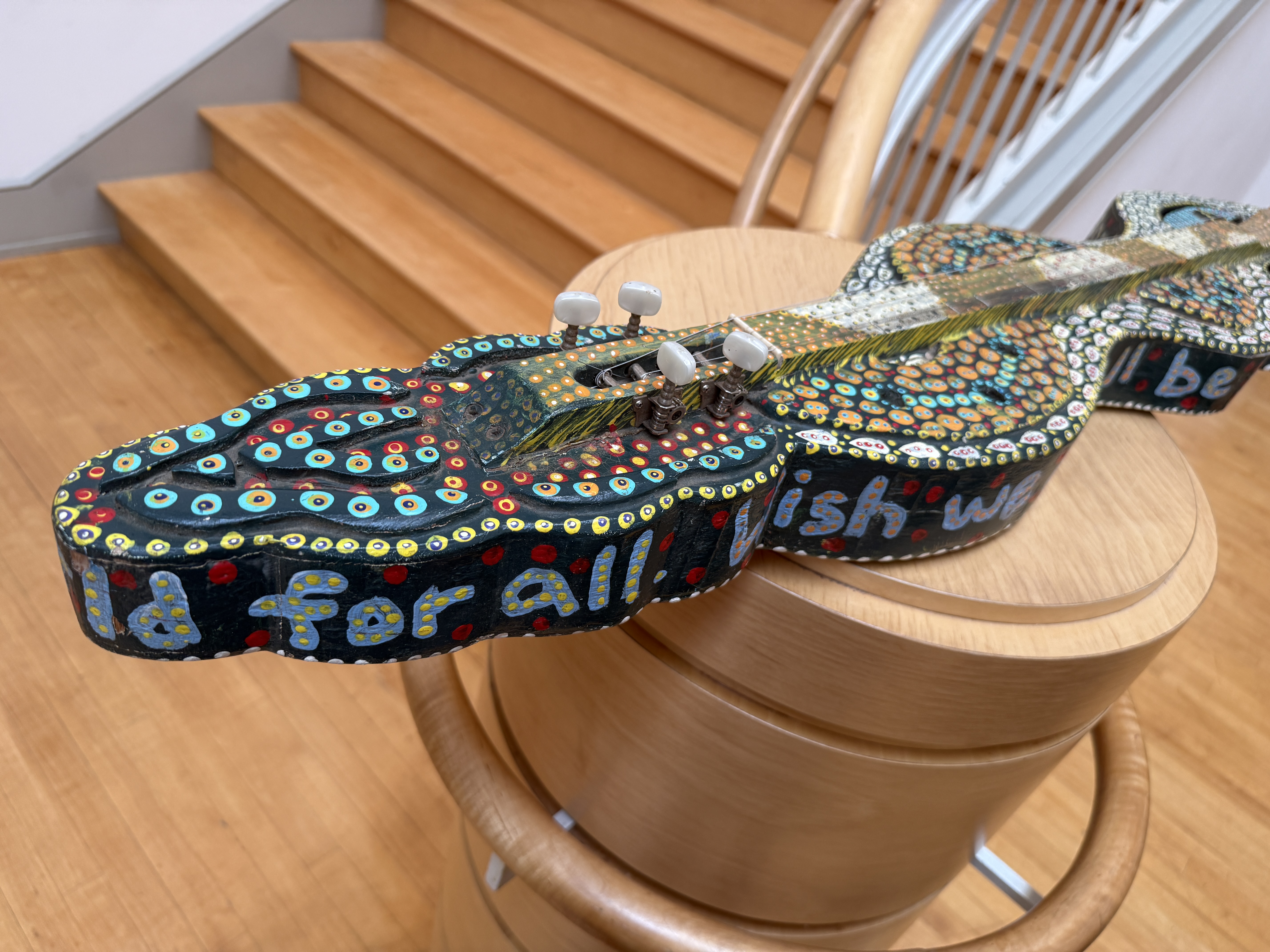 The snake dulcimer, painted with dense dot-work patterns and the words "we could all be dazed" along its body, displayed on a wooden pedestal