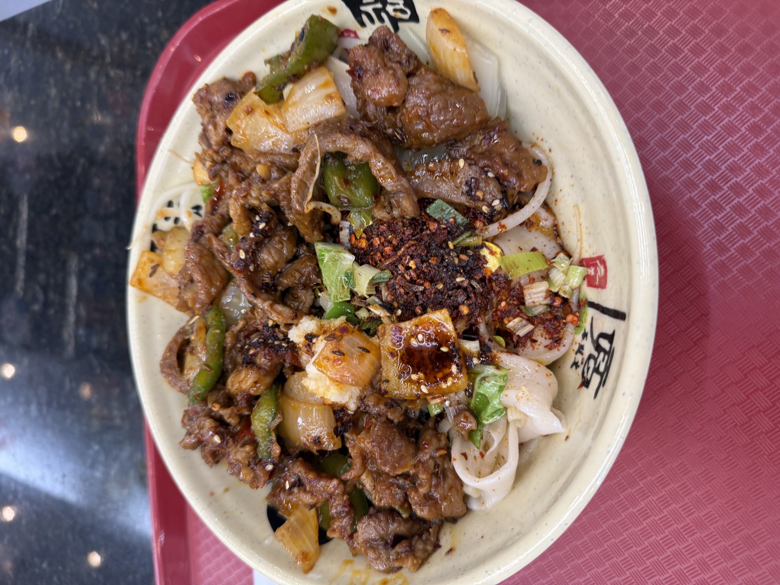 Overhead view of a generous bowl of biang biang noodles from Xi'an Gourmet House, served on a red plastic tray. The wide, flat hand-pulled wheat noodles peek out from beneath an abundant topping of stir-fried lamb in a glossy dark sauce, mixed with chunks of white onion, sliced green bell pepper, and bright green leaves of leafy vegetable. A heaping pile of red chili flakes and toasted sesame seeds sits in the center, with a small drizzle of red chili oil pooled around a piece of onion. Scattered scallion greens add bright color throughout. The cream-colored ceramic bowl is stamped with red Chinese calligraphy characters.