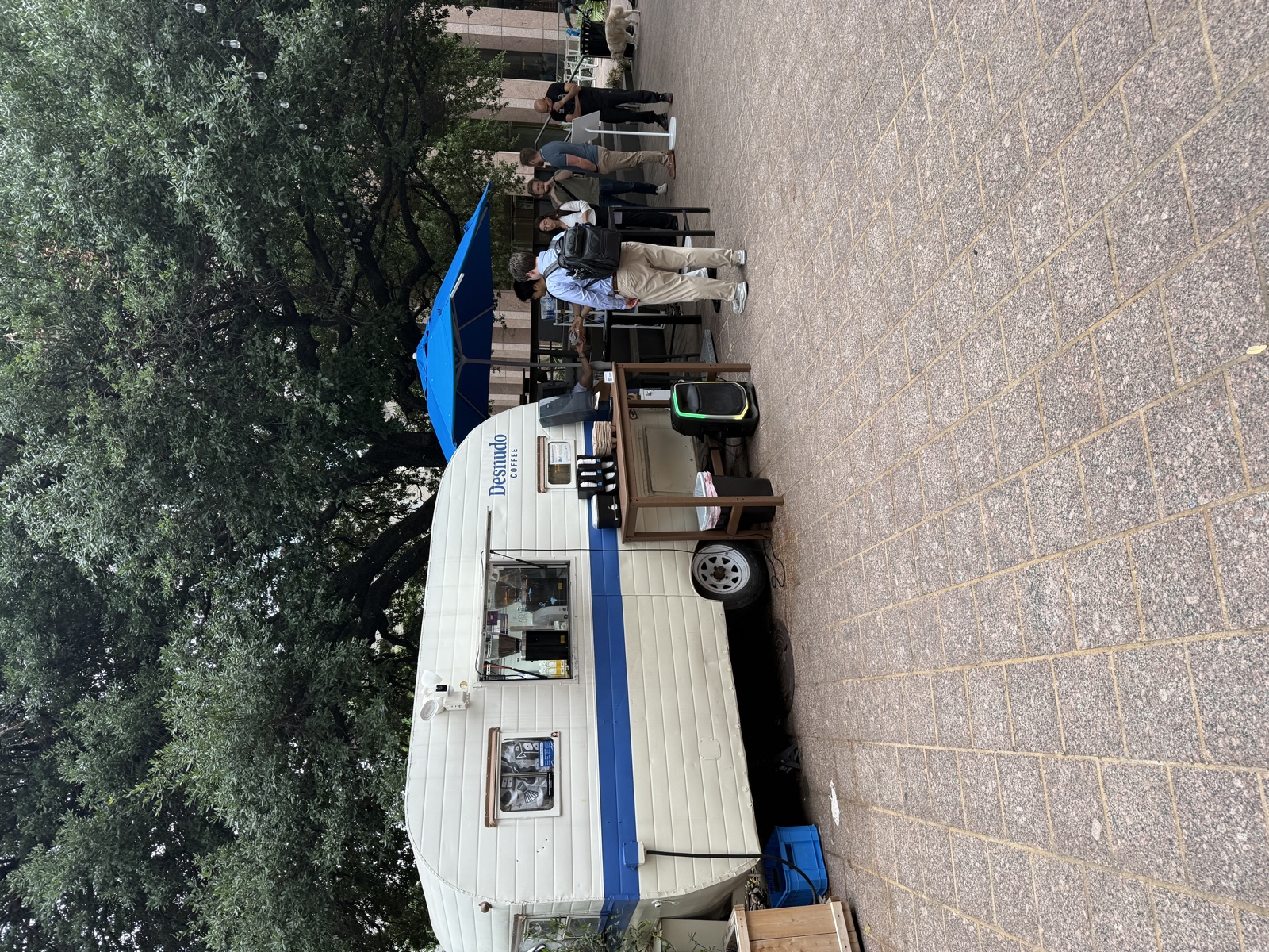 A small vintage white camper trailer with a horizontal blue stripe along its lower half and the word 'Desnudo Coffee' painted in blue near the back, parked on a brick plaza beneath the spreading branches of a massive live oak. The serving window is open, with coffee equipment visible inside. A wooden side table holds three coffee dispensers, and a larger metal-topped service table sits next to the trailer with espresso machines and a tall blue umbrella overhead. A small line of customers stands waiting their turn, dressed in business-casual clothing. A string of cafe lights runs through the trees overhead, and a fluffy white dog can be seen at the right edge of the frame near the building.