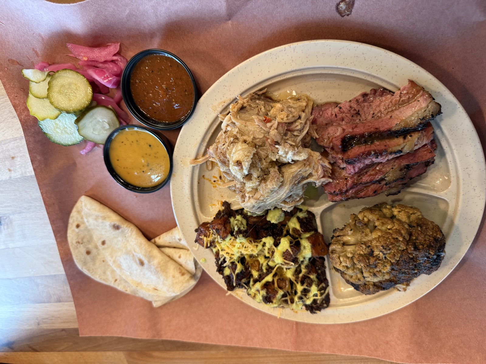 An overhead view of a barbecue plate at Leroy and Lewis, served on a divided cream-colored melamine plate set on butcher paper. The top-left compartment holds a charred whole head of smoked cauliflower. The top-right compartment holds chunks of dark, crusty meat topped with melted yellow cheese and shredded cheddar. The bottom compartment holds three thick slices of pink-rimmed tri-tip on the left and a generous mound of pulled pork on the right. To the right of the plate, a folded flour tortilla, two small black plastic cups holding a yellow mustard-based sauce and a dark reddish-brown sauce, and a pile of cucumber slices and pink-tinged onions in brine.