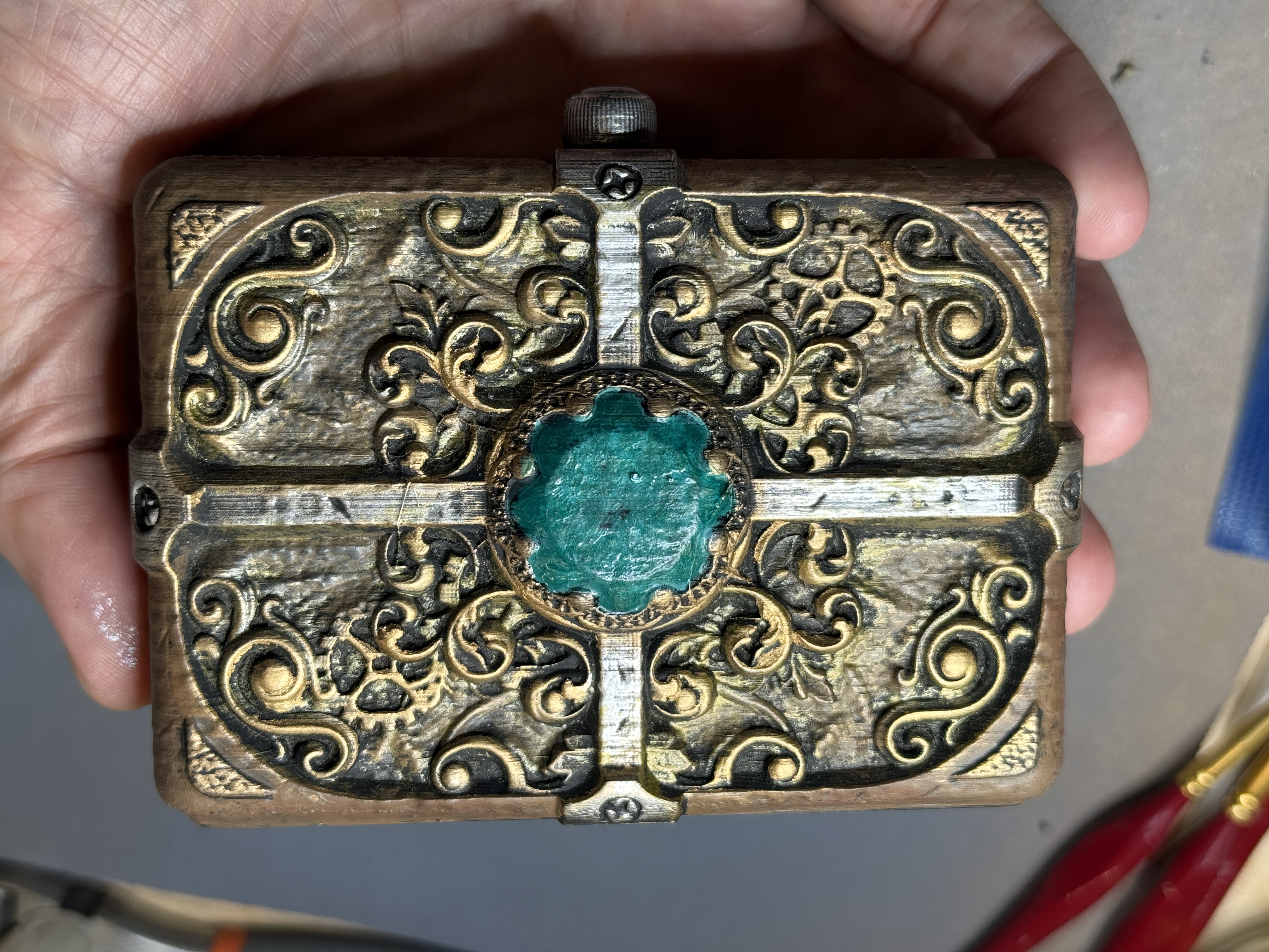 A hand-painted 3D-printed wallet box with ornate gold scrollwork, steampunk gears, and a green gem in the center, held in one hand
